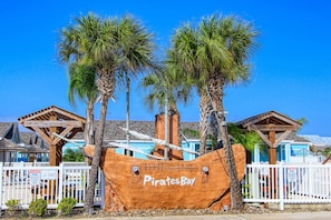 Un bateau pirate en bois, portant l’inscription « Pirates Bay », est amarré devant un bâtiment avec un toit bleu et une clôture blanche.