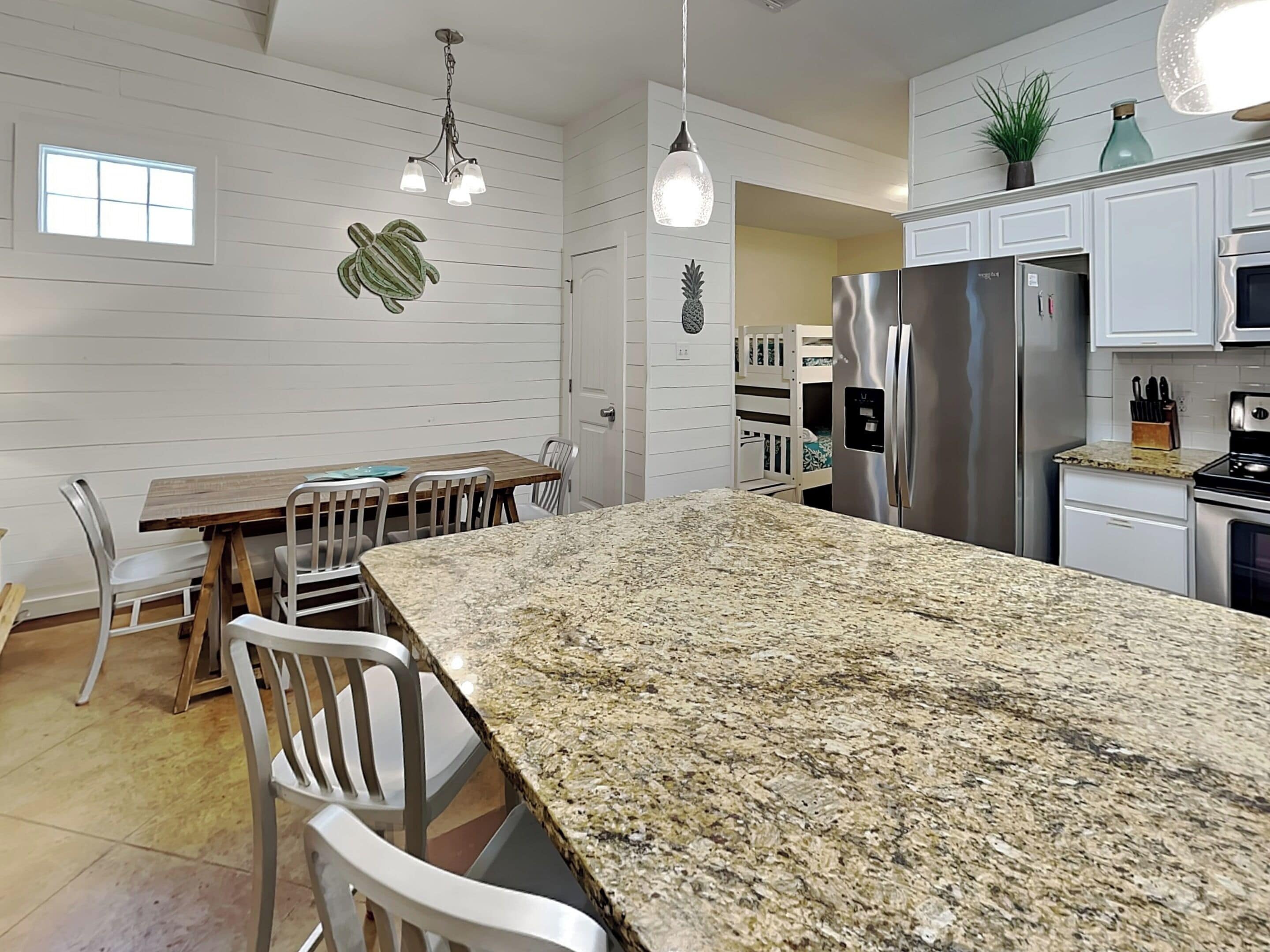 A modern kitchen with white cabinetry, a granite countertop, stainless steel appliances, and a dining area with white chairs.