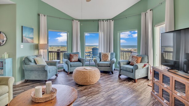 A living room with teal walls, a ceiling fan, a TV, and a large window with a view of the ocean. - Crandall's Cottage by Silver Sands Vacation Rentals (Port Aransas)