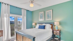 A bedroom with a bed, two bedside tables with lamps, a ceiling fan, and a view through a glass door. - Crandall's Cottage by Silver Sands Vacation Rentals (Port Aransas)