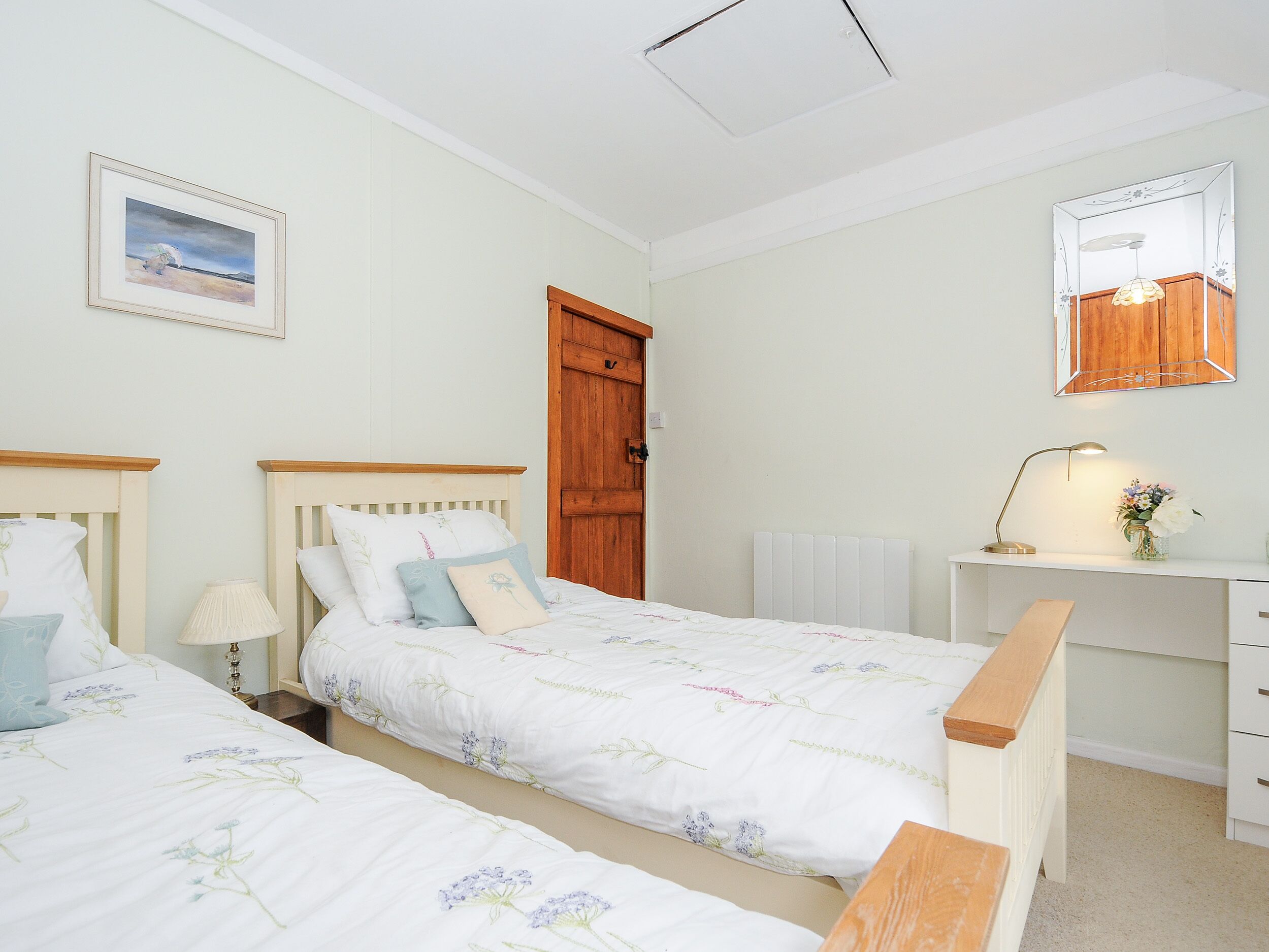 A bedroom with two single beds, a wooden door, a desk with a lamp, and a mirror on the wall.