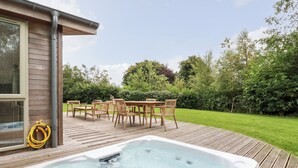 A hot tub on a wooden deck with a table and chairs, surrounded by a grassy yard and trees. - 14 Horizon View (Liskeard)