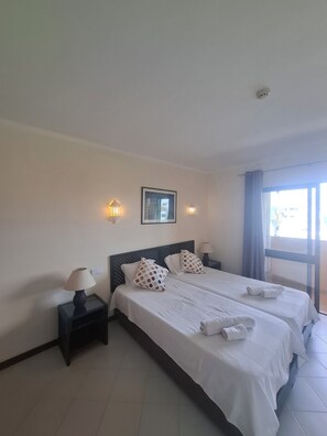 A hotel room with two beds, a nightstand with a lamp, wall sconces, a framed picture, and a window with curtains.
