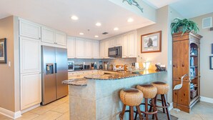 Una cocina moderna con armarios blancos, una isla de granito con taburetes altos, electrodomésticos de acero inoxidable y una estantería de madera.