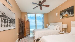 Un dormitorio con dos camas, un ventilador de techo, un ventanal con vistas a la playa y un cuadro de una concha.