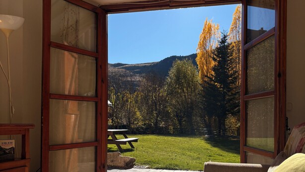 Ein offener Raum mit Holztisch und einer Couch, Blick auf einen grünen Rasen mit Bäumen und einem Picknicktisch draußen.
