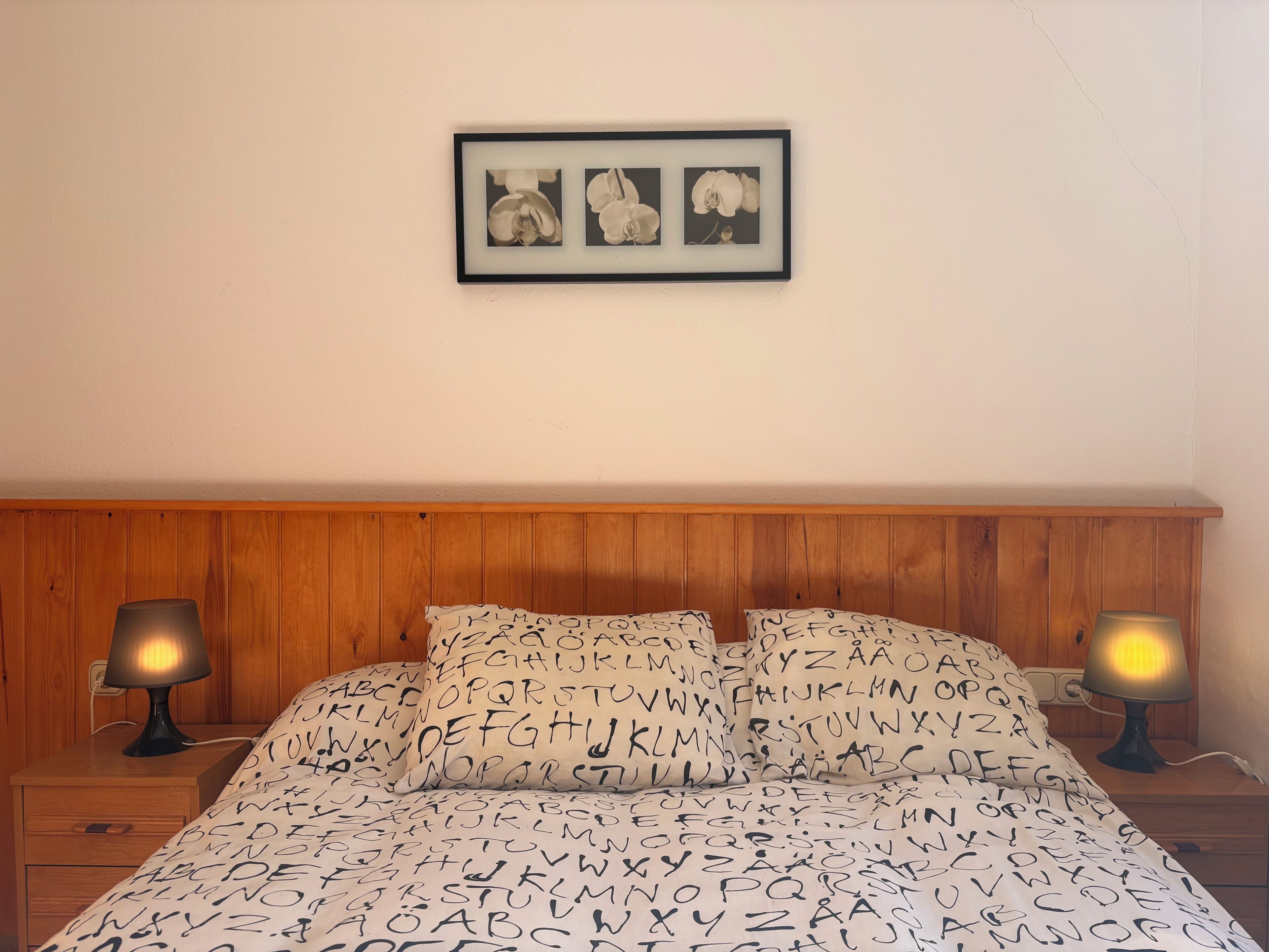Un dormitorio con cabecera de madera, cama con ropa de cama de diseño alfabético, dos lámparas de noche y un cuadro enmarcado sobre la cama.