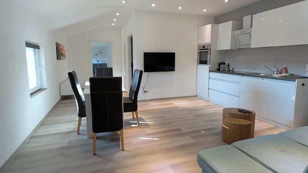 A modern kitchen with white cabinets, a built-in oven, and a microwave. There is a dining area with a black table and chairs, and a TV mounted on the wall.