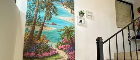 Una escalera con baldosas color beige, barandilla de metal negro y un cuadro grande con paisaje tropical en la pared.