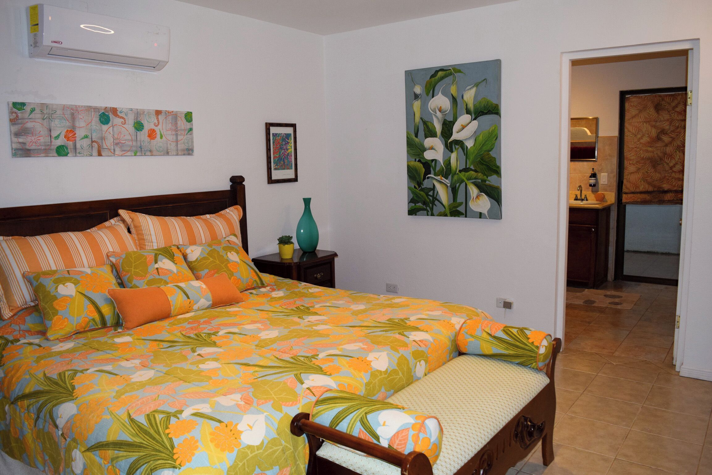 Un dormitorio con edredón floral, cabecera de madera, mesita de noche con un florero y un cuadro de flores en la pared.