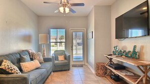 A living room with a grey sofa, a ceiling fan, a flat-screen TV, and a wooden coffee table with a 'Beach' sign. - Family Tides Beach House by Silver Sands Vacation Rentals (Port Aransas)