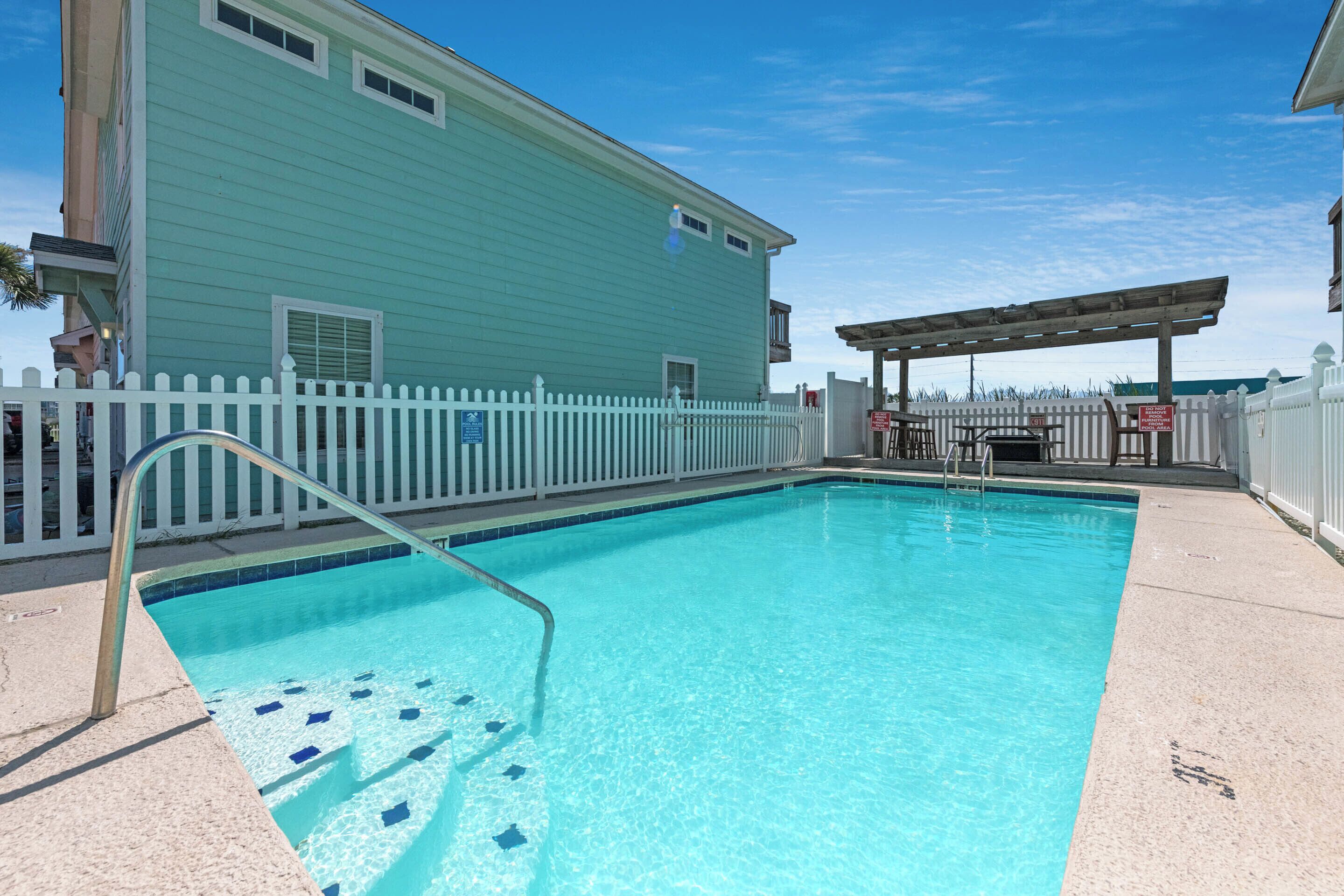 A swimming pool with a white picket fence, a covered patio area, and a light blue building with a balcony.