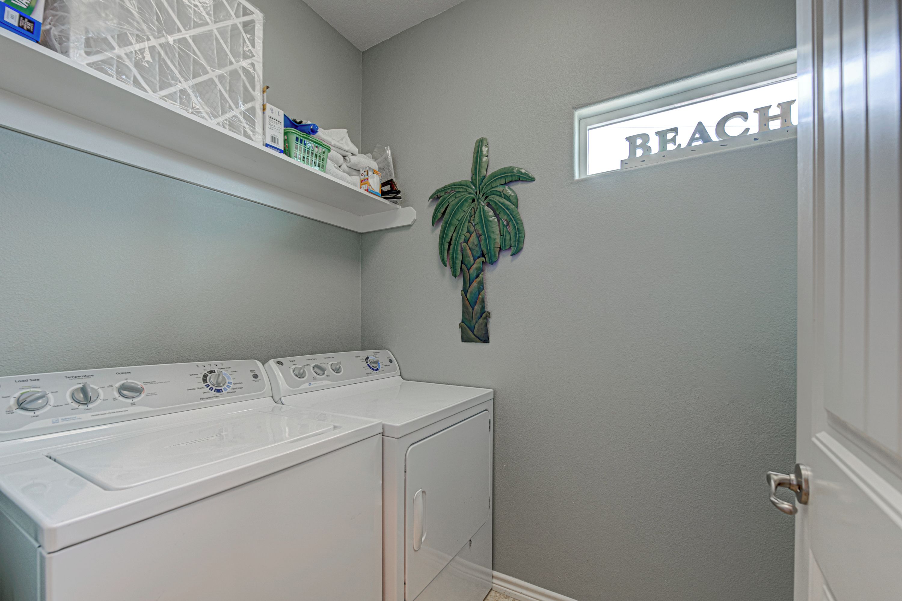 A laundry room with a white washer and dryer, a shelf above the dryer, a wall-mounted plant, and a 'BEACH' sign on the door.