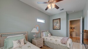 Une chambre avec deux lits, un ventilateur de plafond, un tableau encadré et une petite fenêtre.