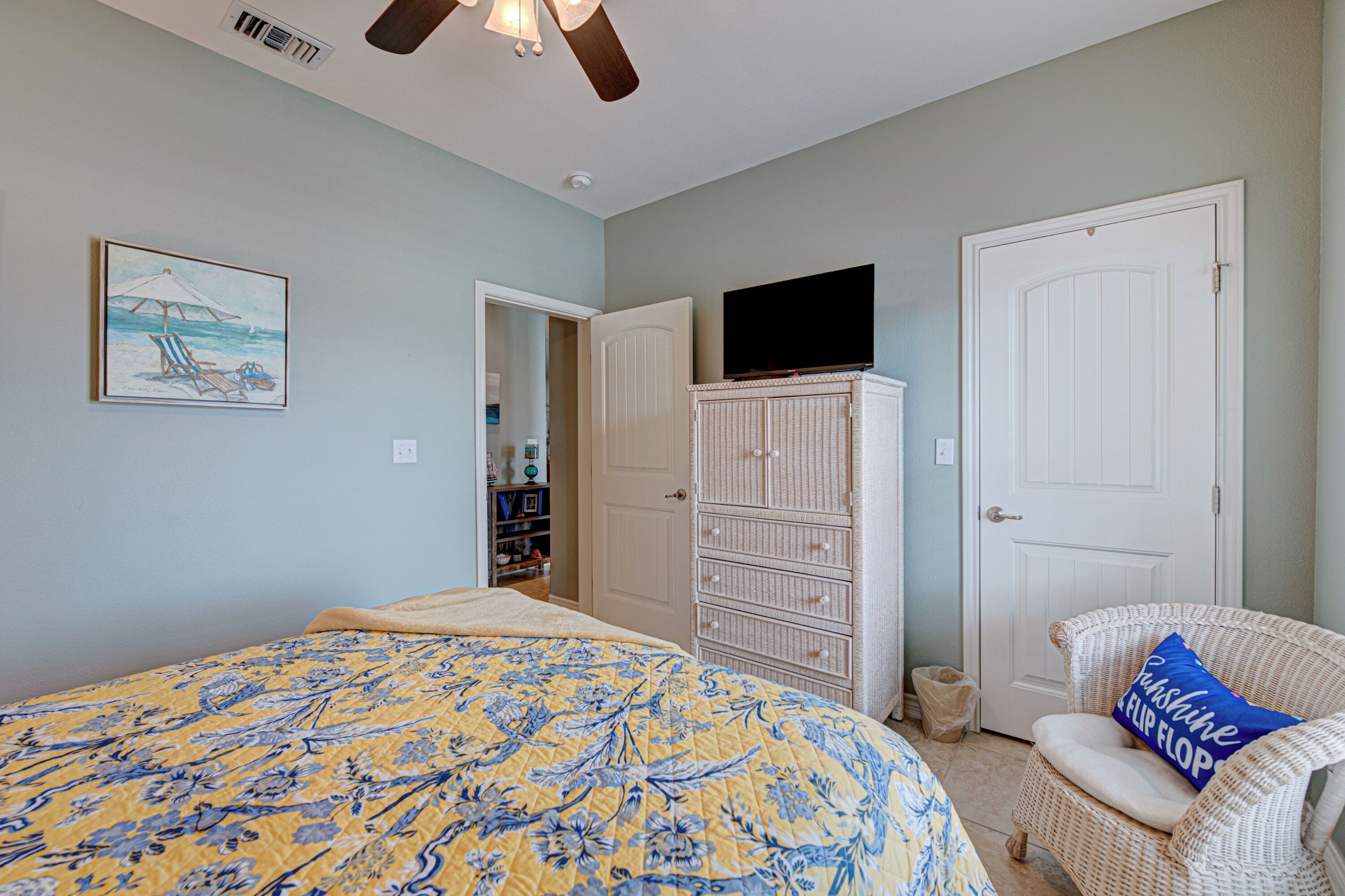 A bedroom with a bed, a television on a dresser, a wicker chair, and a door.