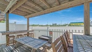 Une terrasse en bois à l’extérieur, avec des tables de pique-nique et des bancs, une clôture blanche en bois et un ciel bleu dégagé.