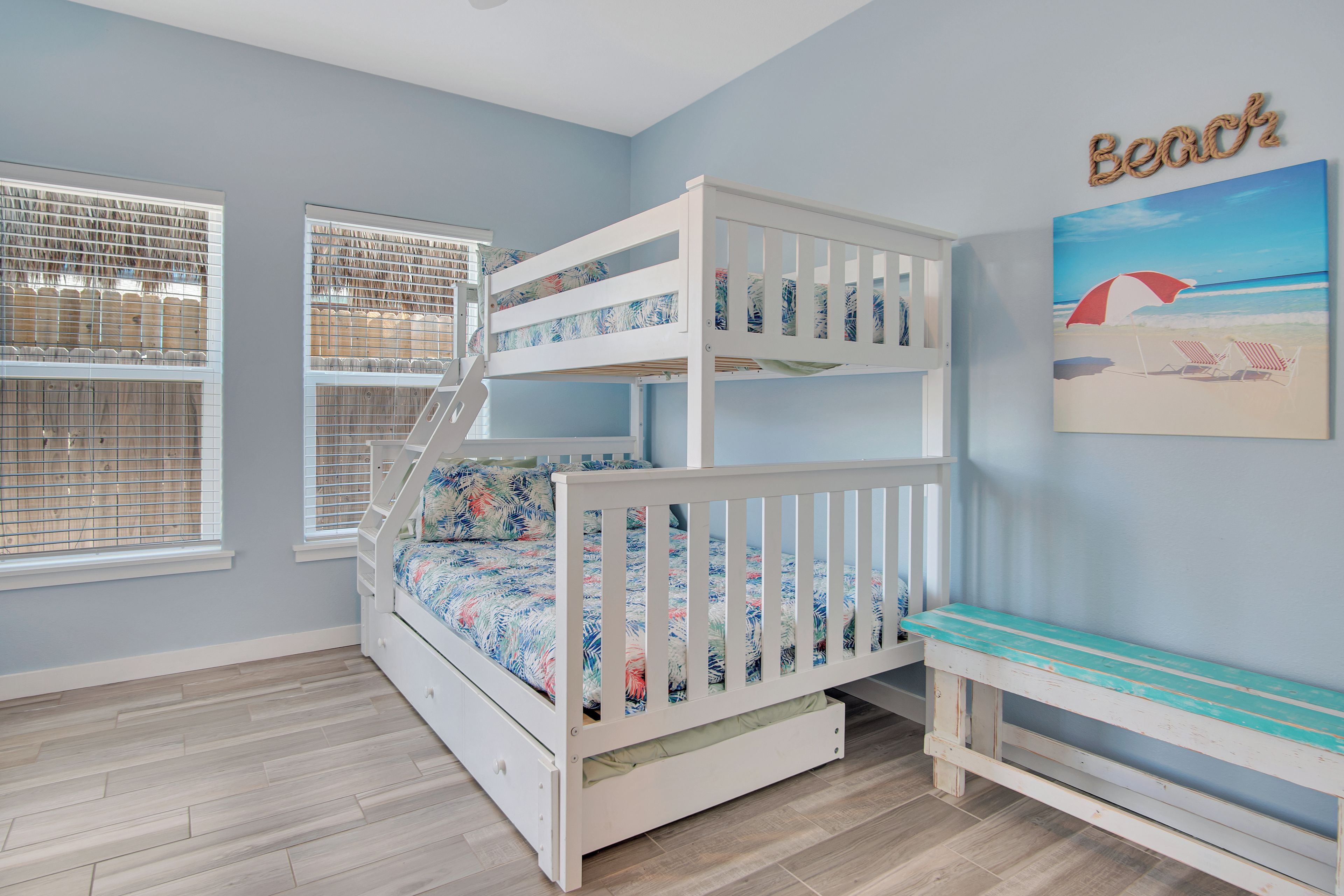 A bunk bed with a floral bedspread, a bench, and a beach-themed wall art.