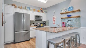 A kitchen with white cabinets, stainless steel appliances, a granite countertop, and a wooden bar stool area. - Fin And Tonic by Silver Sands Vacation Rentals (Port Aransas)