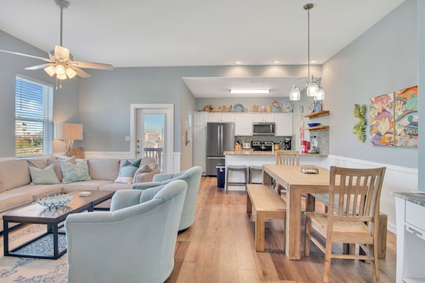 A modern kitchen with white cabinets, a stainless steel refrigerator, and a microwave. There is a dining area with a wooden table and chairs. - Fin And Tonic by Silver Sands Vacation Rentals (Port Aransas)