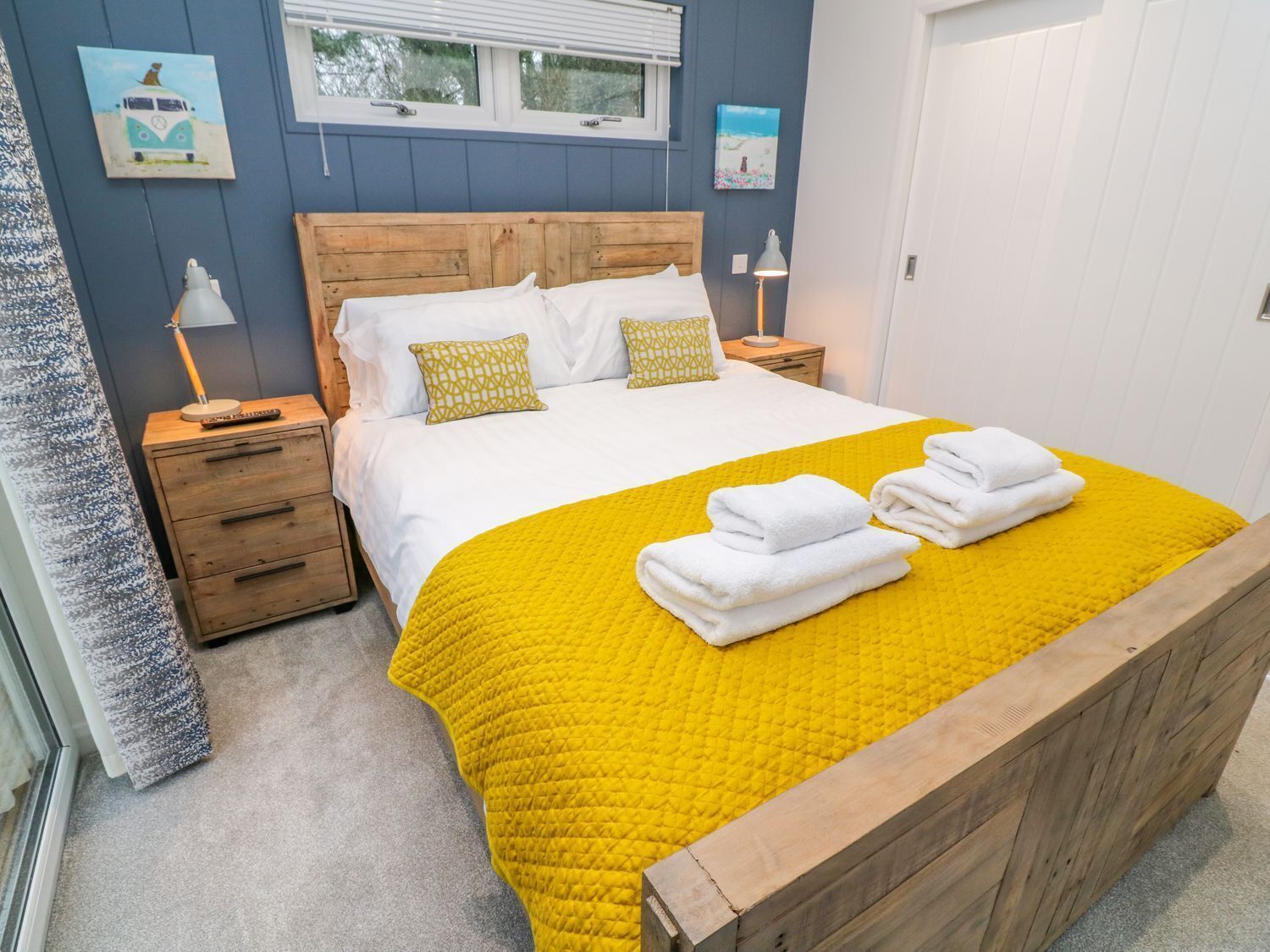 A neatly made bed with a yellow quilt, white pillows, and a wooden headboard. There are two bedside tables with lamps, and artwork on the walls.