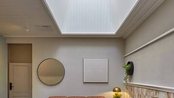Un dormitorio con tragaluces, una cama, una zona de estar con sofás de cuero, un espejo redondo, un teléfono en la pared y una mesita con una lámpara.
