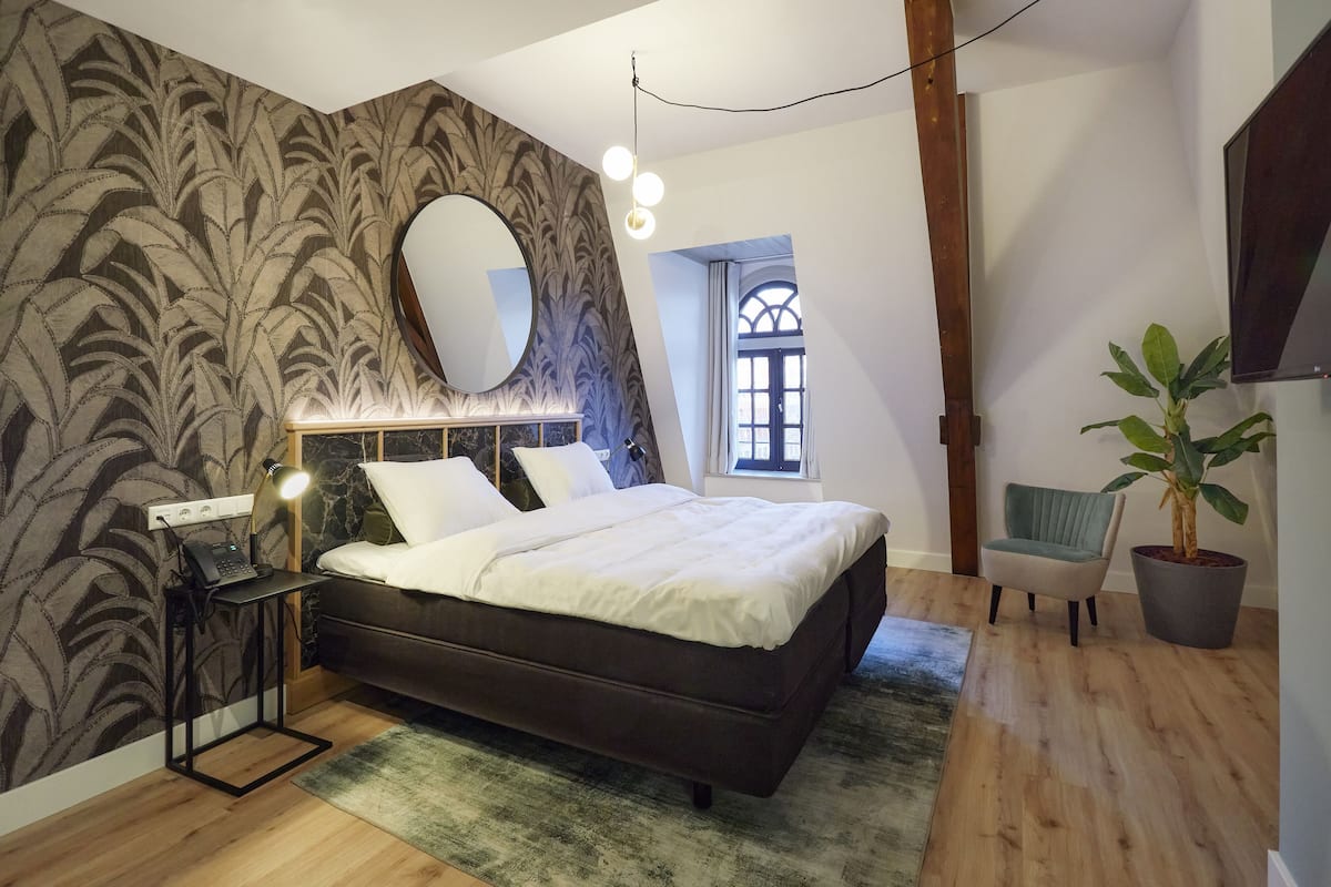 A bedroom with a bed, a bedside lamp, a mirror, a potted plant, and a window.