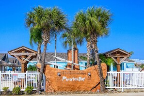 A wooden pirate ship with the name 'Pirates Bay' in front of a building with a blue roof and white fence. - Happiness Comes In Waves by Silver Sands Vacation Rentals (Port Aransas)