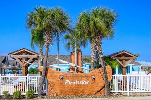 A wooden pirate ship with the name 'Pirates Bay' in front of a building with a blue roof and white fence.