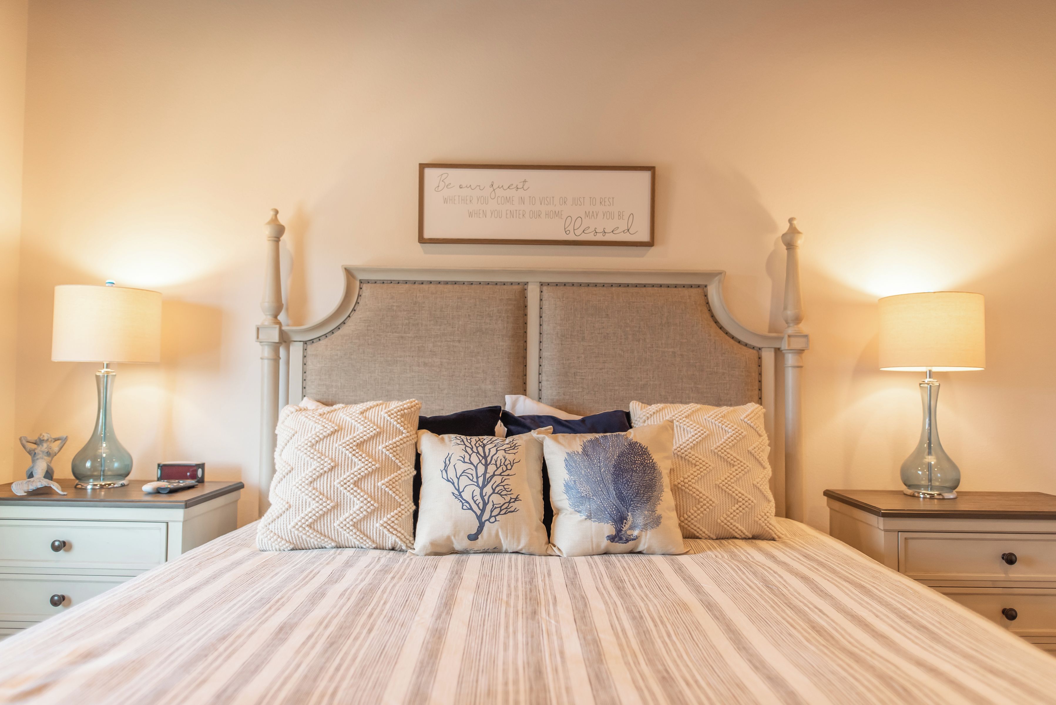 A neatly made bed with a striped bedspread, flanked by two bedside tables with lamps, a framed sign above the bed, and decorative pillows.