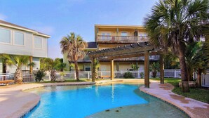 A swimming pool with a wooden deck and palm trees, surrounded by buildings with balconies and a covered patio area. - Getaway by Silver Sands Vacation Rentals (Port Aransas)