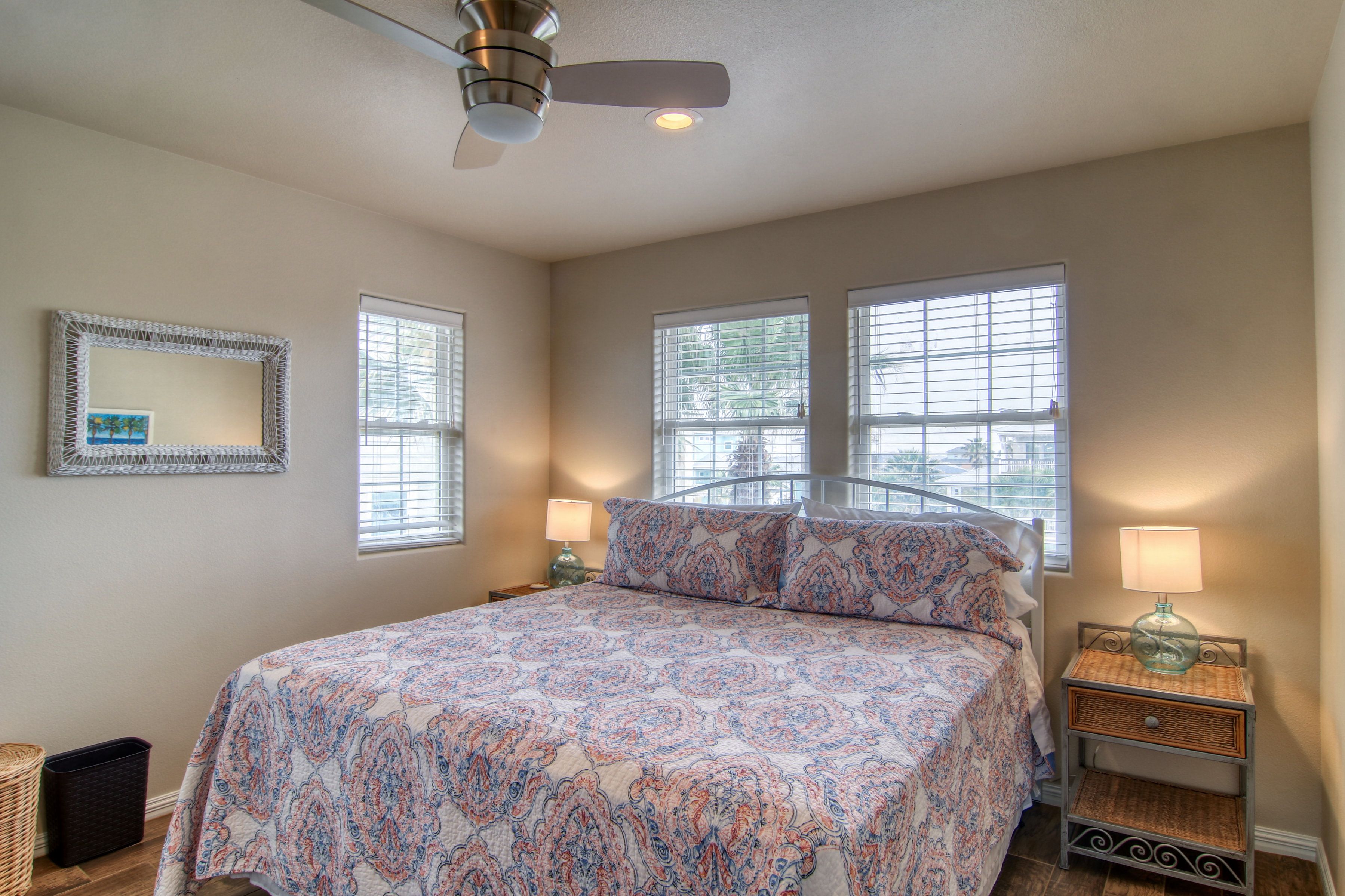 Une chambre à coucher comprenant un lit, deux tables de chevet, une commode, un ventilateur de plafond et deux fenêtres.
