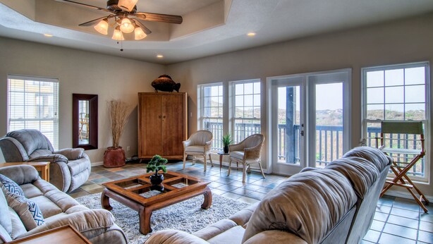 A spacious living room with a high ceiling fan, large windows, and a view of the outdoors. - Happy Heron by Silver Sands Vacation Rentals (Port Aransas)
