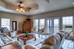 A spacious living room with a high ceiling fan, large windows, and a view of the outdoors.