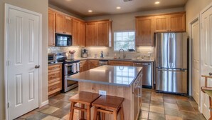 A modern kitchen with wooden cabinets, stainless steel appliances, and a central island with stools. - Happy Heron by Silver Sands Vacation Rentals (Port Aransas)