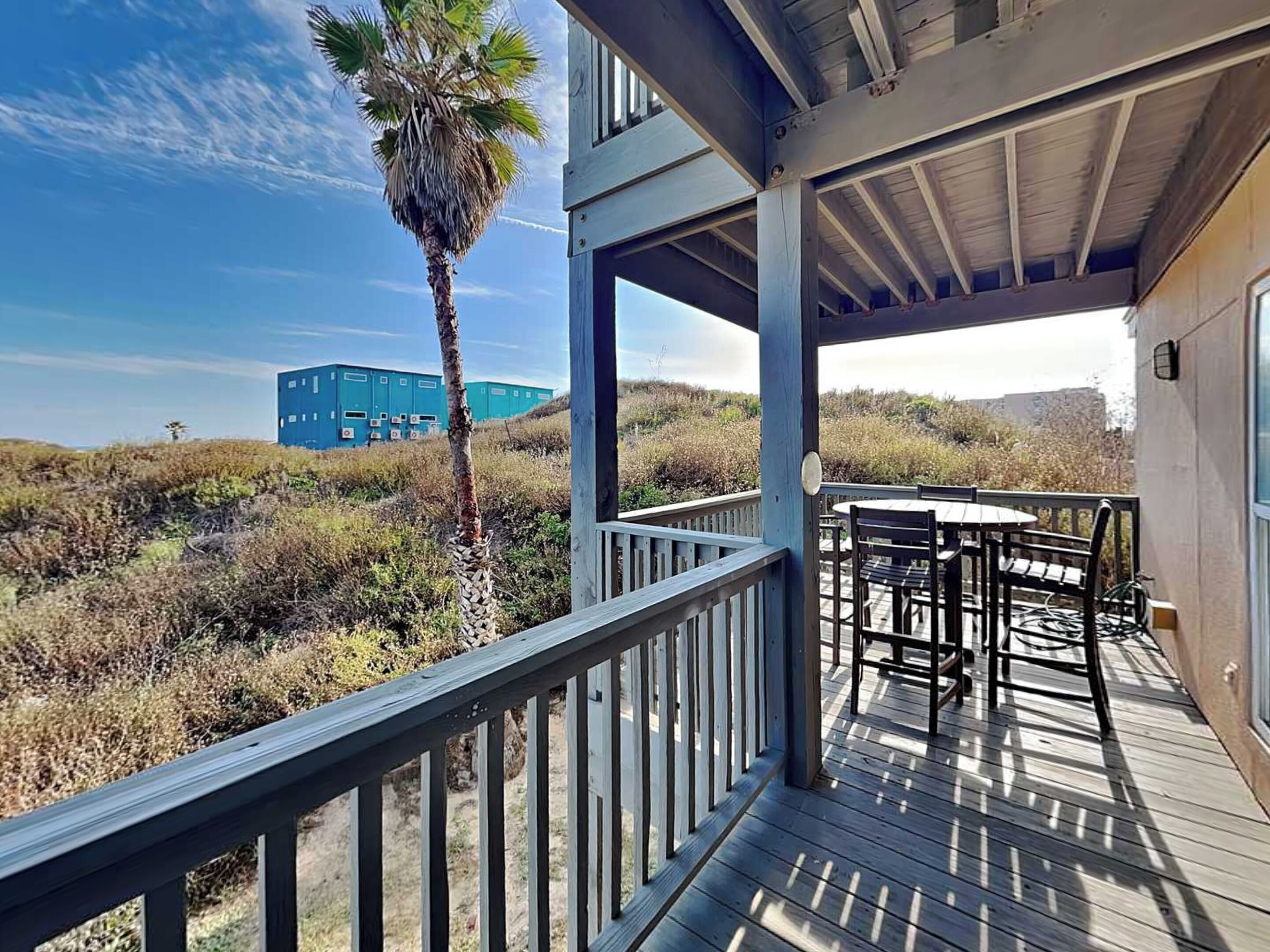A wooden deck with a table and chairs, overlooking a landscape with dry vegetation and a blue building in the distance.