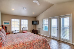 A bedroom with a large bed, a TV, a chair, a table, and a balcony with sliding doors.