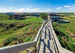 Une promenade en bois mène à une grande maison dotée d’un balcon, qui est entourée de verdure et d’autres maisons.