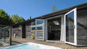 A modern outdoor hot tub area with a wooden deck, a glass-enclosed hot tub, and a sliding glass door leading to a kitchen. - 5 Meadow Retreat (Liskeard)