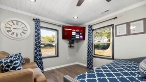 A room with a bed, a couch, a wall clock, a TV mounted on the wall, and two windows with curtains. - Sharpe Tiny House — Cozy, Remodeled Home with Big Amenities (Statesville)