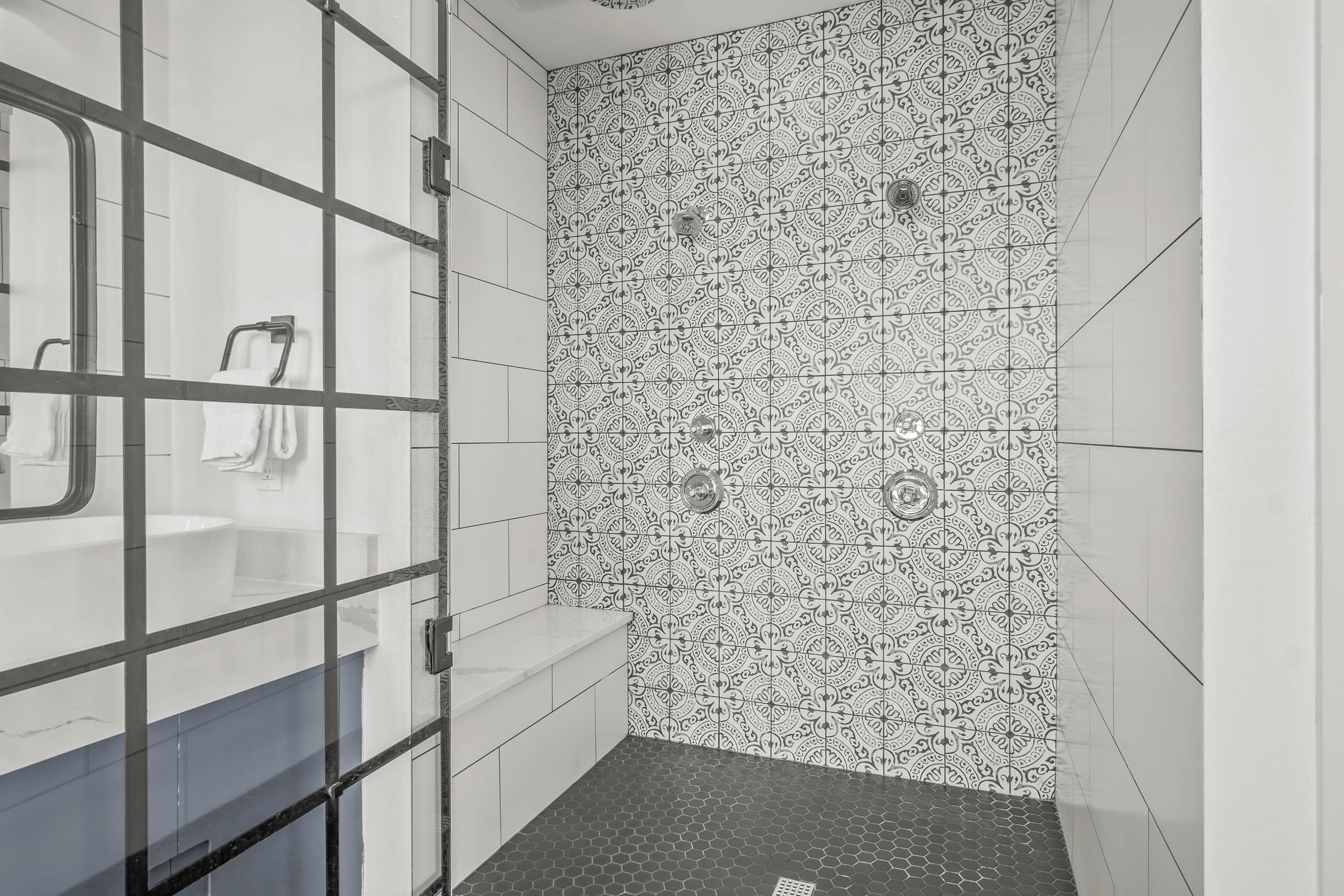 Une salle de bain avec un mur recouvert de carreaux à motifs, une douche en verre et un lavabo doté d’un plan de travail blanc.