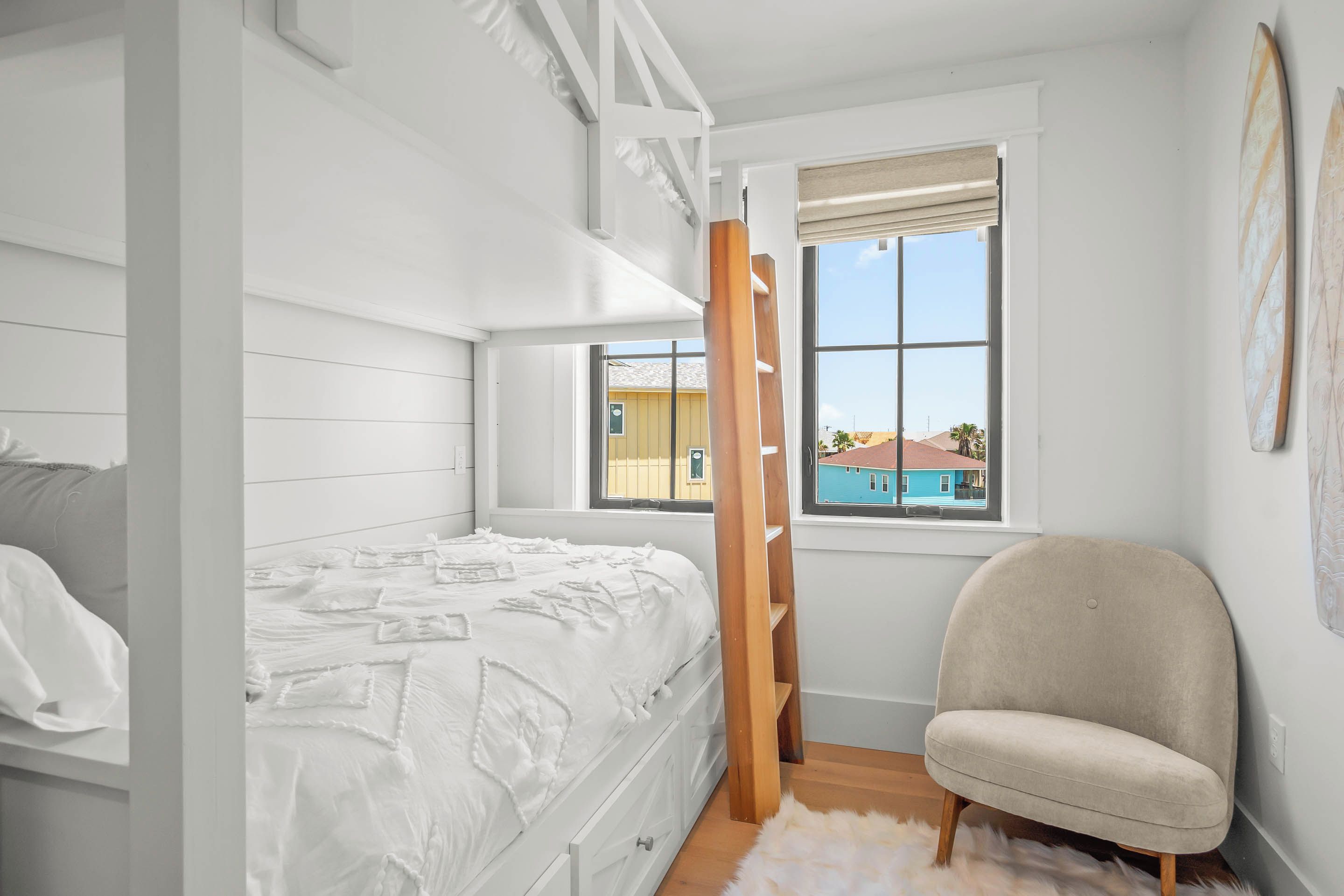 Une chambre avec un lit superposé, une chaise, une fenêtre donnant sur une maison bleue et un tapis moelleux.