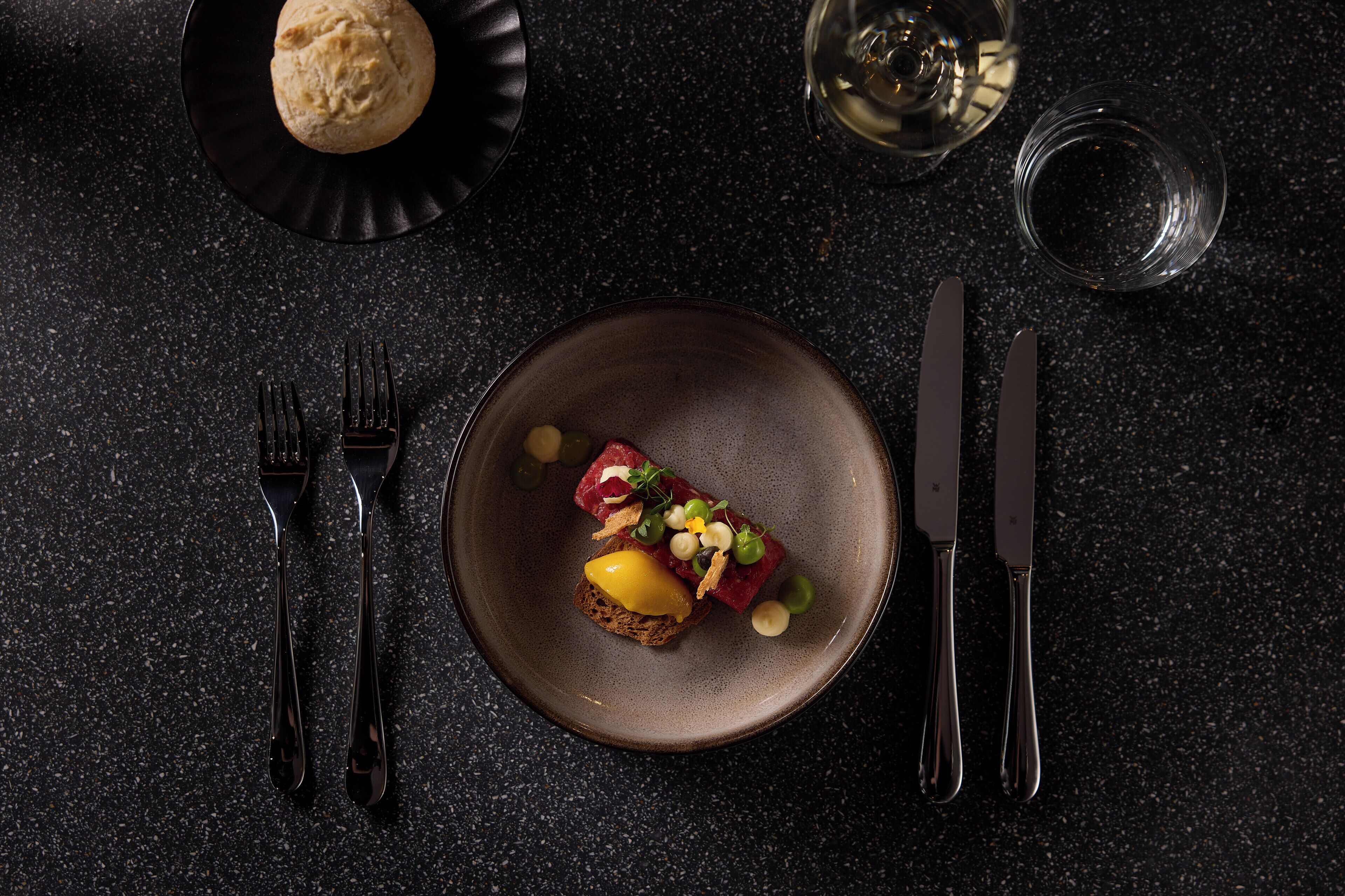 A dark, textured table with a plate of food, a glass of water, and cutlery.