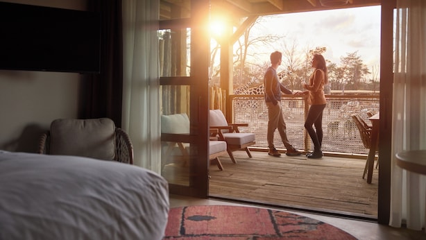 A room with a bed, a sofa, and a view of a giraffe enclosure through a glass door. - Safari Hotel Beekse Bergen (Hilvarenbeek)