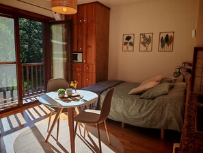 Une pièce avec un lit, une table ornée d’une plante, une chaise et une vue sur l’extérieur.