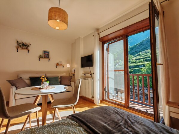 A cozy room with a bed, a round table with chairs, a sofa, a TV, and a large window with a view of green hills.