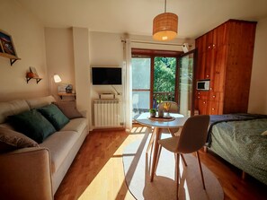 A cozy room with a sofa, a dining table, a bed, a TV, and a view of greenery outside.