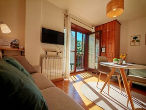 A living room with a sofa, a table with chairs, a TV mounted on the wall, and a window with a view of greenery.