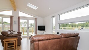 A spacious living room with a brown leather sofa, a wooden coffee table, and a TV on a white stand. Large windows offer a view of the outdoors. - 7 Meadow Retreat (Liskeard)