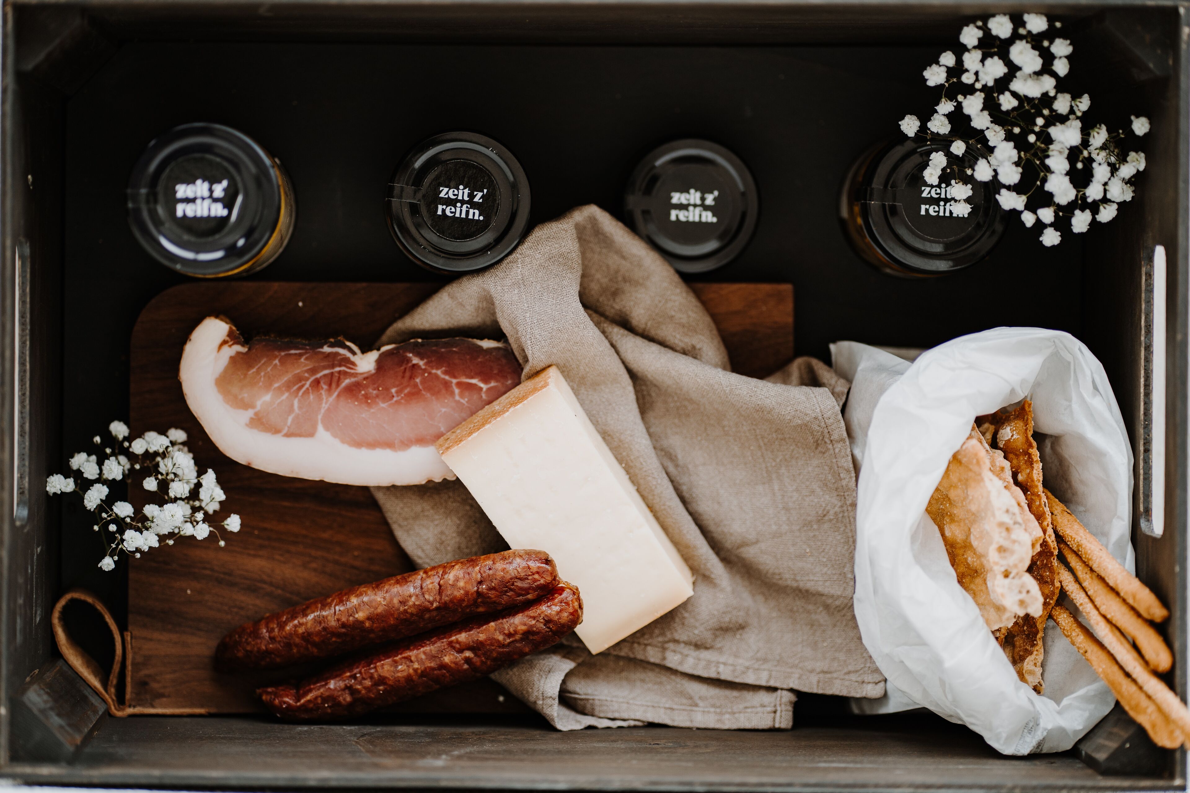 Une planche à découper en bois sur laquelle sont disposés divers aliments, notamment un morceau de viande, du fromage et des saucisses, ainsi qu’une serviette en tissu et un sac en papier contenant des gressins.