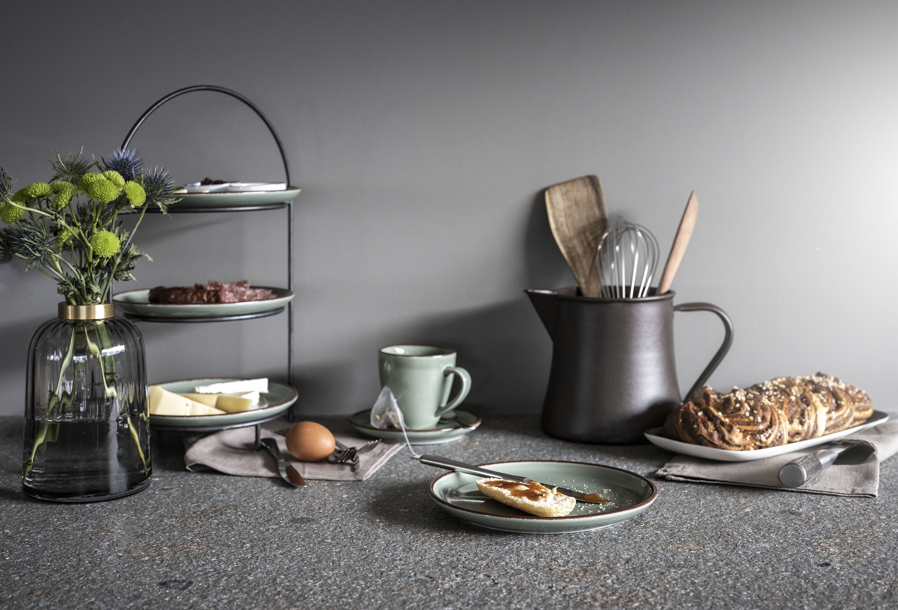 A kitchen counter with a tea set, a plate of pastries, a vase with flowers, and a metal container with utensils.